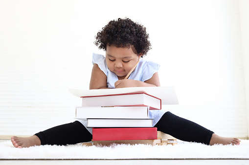 Little African American school girl with curly hair studied and doing homework, reading book and writing notes from a pile of books, kid learning and education at home concept.