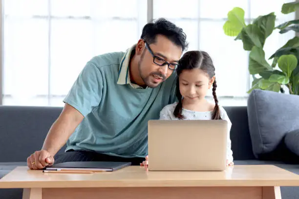 Young handsome father teach adorable girl daughter to do school homework, using laptop computer for e-learning study at home, dad and kid spend time together for education and searching Internet.