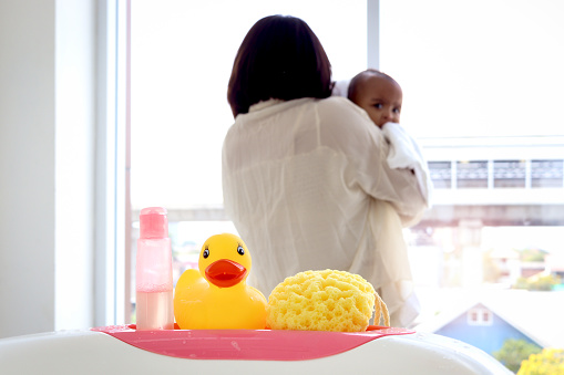 Yellow duck toy, shampoo soap and sponge (baby toiletries) on bathtub in bathroom with blurry background of mother holding infant baby covered with white towel.