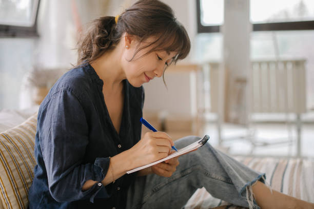 Asian woman making notes in notebook, working at home on bed in cozy bedroom. Concept of freelancer