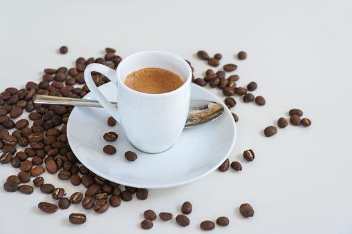 Espresso in a small coffee cup, spoon and roasted beans on a white table, copy space, selected focus, narrow depth of field