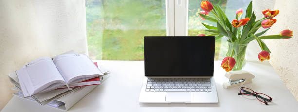 istock Laptop with blank black screen on a white desk in front of the window, also folder, organizer and tulip bouquet, mock up for study or home business, panoramic format, copy space 1391653058