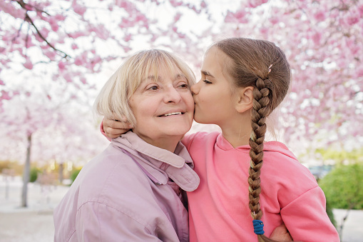 Girl kisses her grandmother near the blooming branch of sakura during walking in the spring garden. Happy spring vibes. Active life in any season, age and weather. Happy seniors in retirement