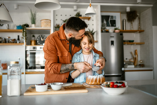 Father and daughter at home making a cake together. Decorating it with powdered sugar and having fun in the kitchen.