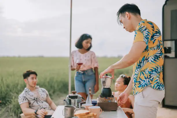 Photo of Asian chinese gay man making coffee and bonding time with friends beside paddy field in front of campervan
