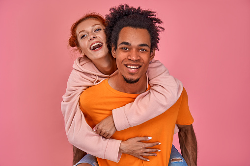 Photo of playful African American man gives piggyback ride to red-haired girlfriend, have fun together, smile happily, pose over pink background. Happy man carries a woman on the back.