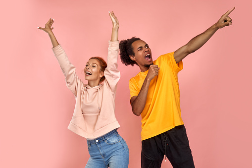 A mixed race couple of young funny and happy dark-skinned man and ginger woman dancing and reading hip-hop at studio on pink trendy color background. Human emotions, youth, love and lifestyle concept