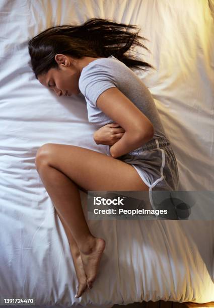 Full Length Shot Of An Attractive Young Woman Lying Alone On Her Bed And Suffering From A Tummy Ache Stock Photo - Download Image Now