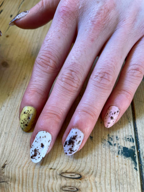 Image of unrecognisable woman's hand on wooden table showing nail varnish manicure design, white, yellow and pink speckled egg design Stock photo showing close-up view of a woman displaying her manicured painted nails designed like speckled Easter eggs. yellow nail polish stock pictures, royalty-free photos & images