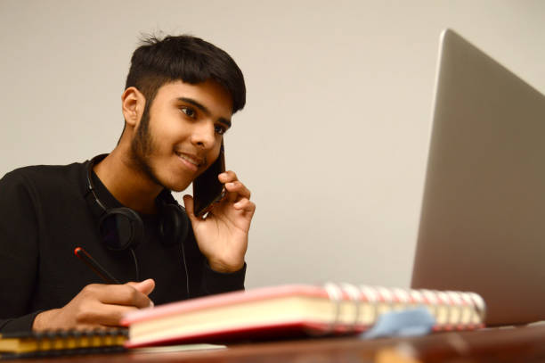 horizontal photograph of  one smiling teenage boy or young male with black hair and beard  looking in the blank while writing and taking notes and talking over mobile phone, laptop screen busy multi tasking with smile on his desk over grey background - multi tasking asian and indian ethnicities asian ethnicity lifestyles imagens e fotografias de stock