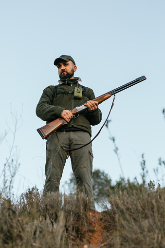 Low angle of bearded male standing with rifle and observing environment while looking for prey on rural terrain