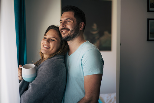Young heterosexual couple standing behind the window and smiling. They are looking through the window. She is holding cup of coffee.