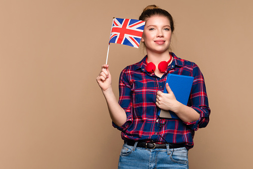 Attractive Student girl holds small UK flag and notebook in studio on beige background. Study abroad concept. International student exchange program. Learning British English with native speaker.