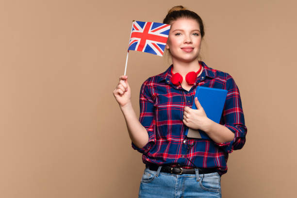 girl holds small UK flag and notebook Attractive Student girl holds small UK flag and notebook in studio on beige background. Study abroad concept. International student exchange program. Learning British English with native speaker. english culture stock pictures, royalty-free photos & images