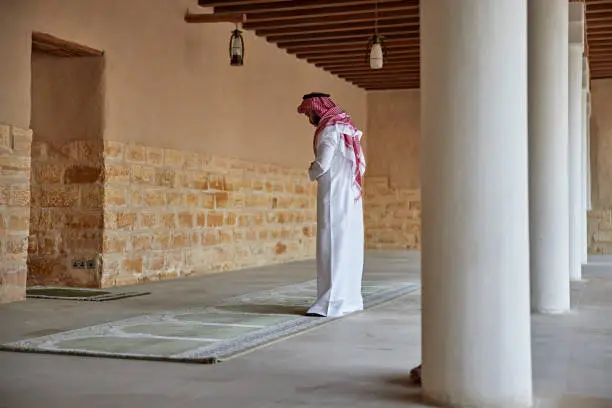 Three-quarter rear view of early 30s Muslim man in traditional attire performing ritual prayer during visit to At-Turaif open air museum near Riyadh. Property release attached.