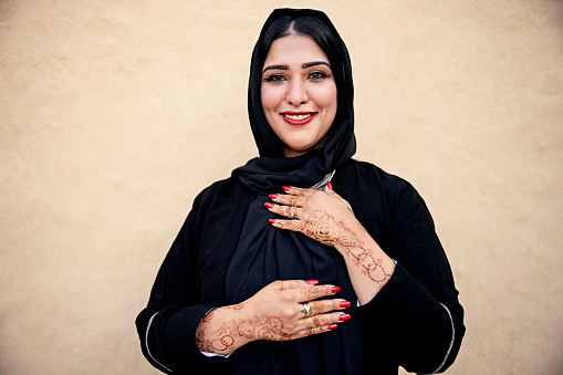 Waist-up view of late 30s woman in traditional black abaya and hijab, displaying her decorated hands, and smiling at camera while standing against textured earthen wall.