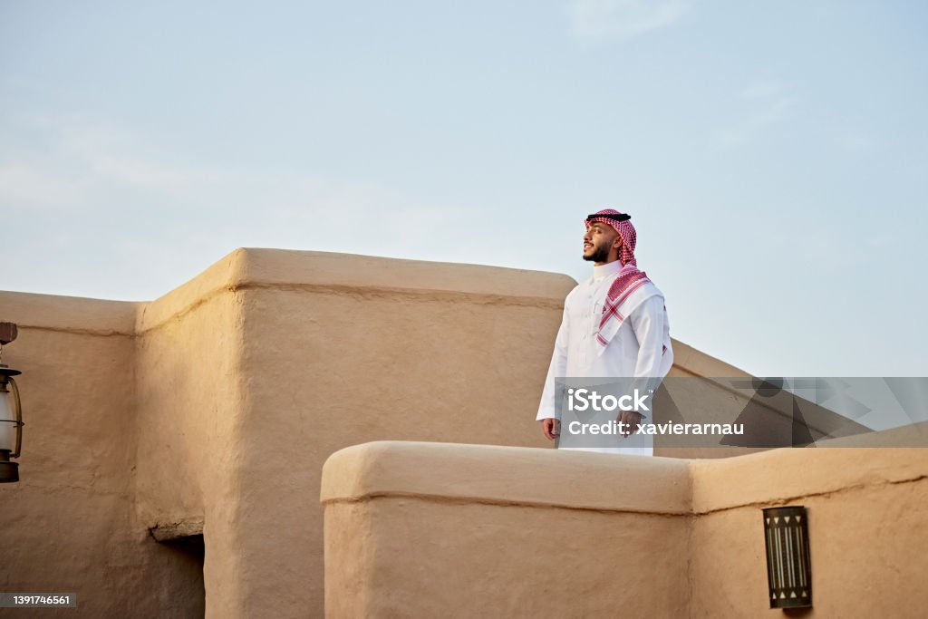 Mid adult Saudi man standing outdoors amidst At-Turaif ruins Waist-up view of bearded Middle Eastern man in traditional attire at UNESCO World Heritage Site near Riyadh and looking away from camera. Property release attached. Saudi Arabia Stock Photo