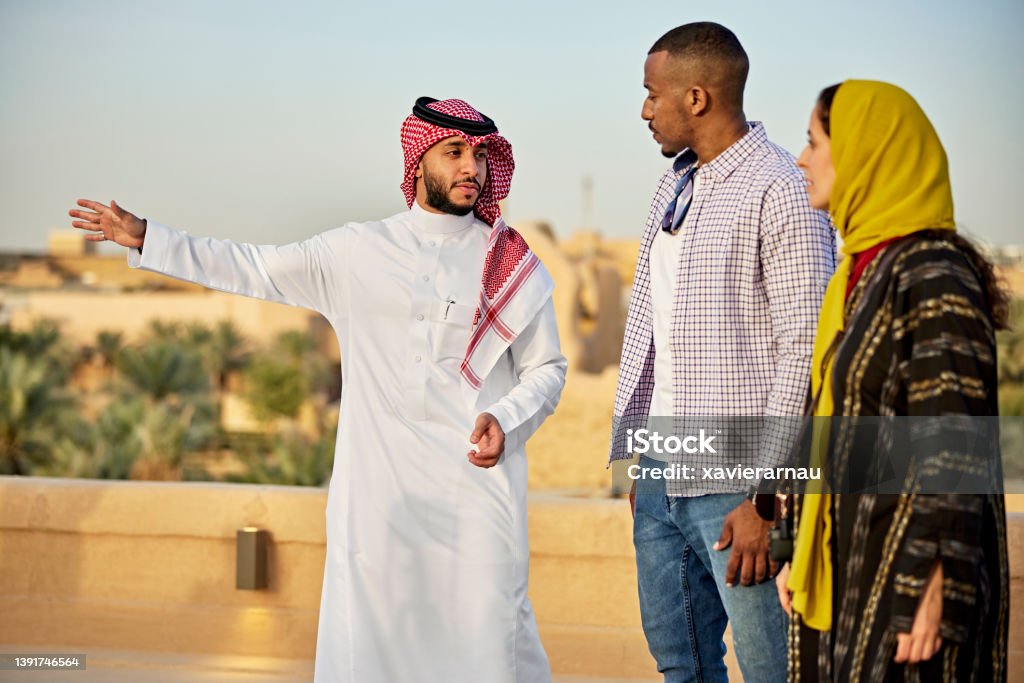 Couple listening to guide as they explore Diriyah ruins Early 30s Middle Eastern man in traditional attire gesturing while describing history of At-Turaif, Saudi Arabia’s first capital, to male and female tourists. Property release attached. Saudi Arabia Stock Photo