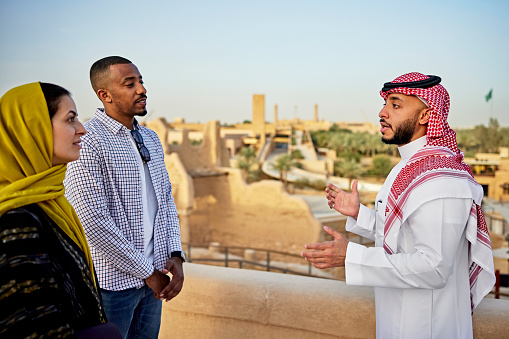 Mid adult couple listening to Saudi man in traditional attire describe preserved ruins from 18th-century site of nation’s first capital, near Riyadh. Property release attached.