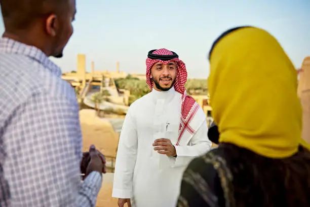 Over the shoulder view with focus on smiling Middle Eastern man in traditional attire discussing history of At-Turaif, original home of Saudi royal family. Property release attached.