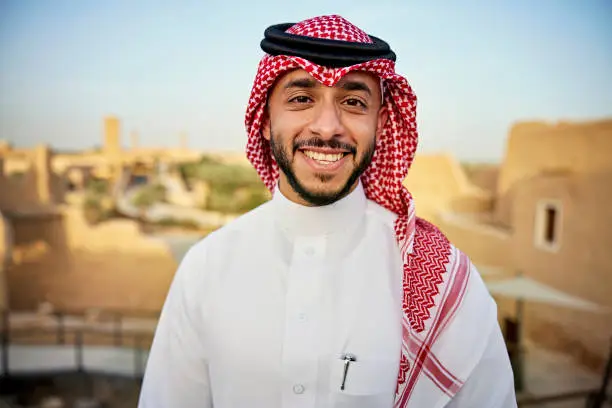 Head and shoulders view of bearded Middle Eastern man in early 30s wearing traditional dish dash, kaffiyeh, and agal and smiling at camera. Property release attached.