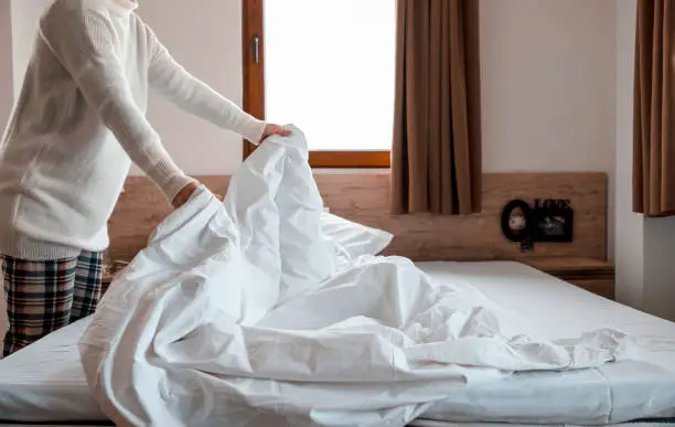 Woman making bed in bright bedroom. Maid Cleaning messy hotel bed cushion and duvet, changing bed sheets with clean. Selective focus. Copy space