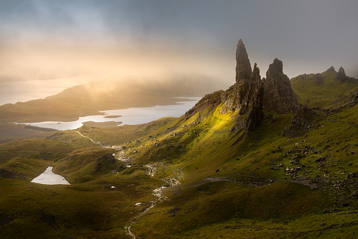 A dramatic view of an iconic Scottish landmark; The Old Man of Storr on The Isle of Skye. The infamous rock pinnacles can be seen poking out of the dark and moody low clouds, as a patch of morning sunlight illuminates the landscape.
