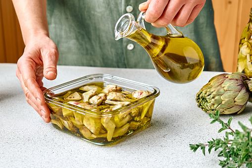 Artichoke hearts marinated with olive oil and herbs. Woman pouring oil in glass jar with pickled artichokes. Homemade healthy eating.
