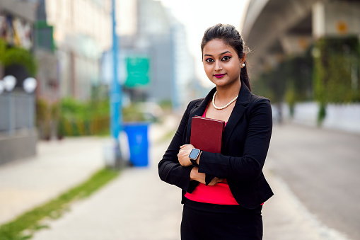 Outdoor portrait of a young teenager woman walking with textbook.She is wearing casual red and black dress and holding diary in her hand.