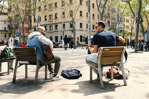 Barcelona, Spain - April 12, 2022: Guitar duo sitting on individual benches play acoustic song while looking at each other. On a hot spring day at noon