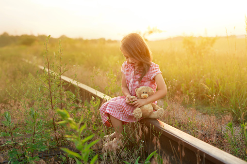 A little girl in a dress sits on an abandoned railroad tracks with a teddy bear at sunset. a lonely child is lost and a toy is his only friend