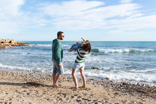 Mother and father playing with their little daughter at the beach. Mother is holding her toddler while is kissing and tickling her, and father is looking at them. Love and laughing at the beach.