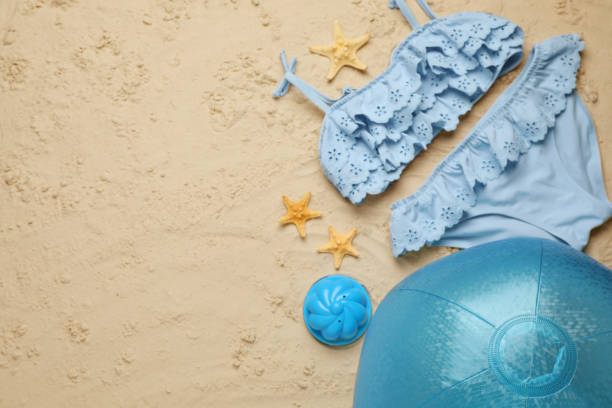 水色のビーチボール、おもちゃ、ヒトデ、砂の上の水着、フラットレイ。テキスト用のスペース
