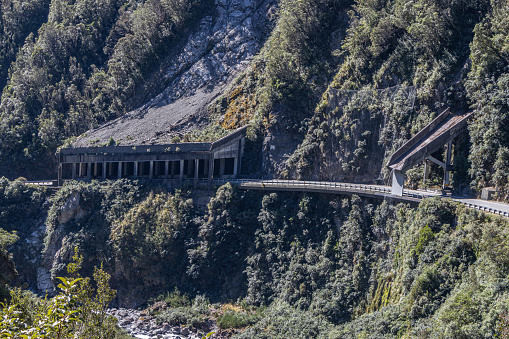 A large structure covers the road at Arthur's Pass, New Zealand, to protect traffic from rock falls. A chute directs water-flow over the traffic to the edge of the gorge. The roof of the shelter is covered in fine debris and remains of trees, and a large rock formation rises behind the shelter. There is a section of wire attached to the hillside between the shelter and the chute, to protect the road from rocks falling. The road runs along the edge of a steep drop into the gorge.