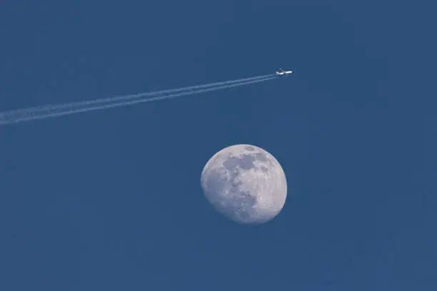 Closeup of the waxing moon and an airplane passing close by, leaving a contrail  - early evening capture against dark blue sky. Stuttgart, Germany April, 13, 2022