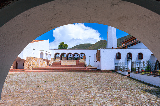 Guatavita, Colombia - The view from under an arched entrance to the main town square in Guatavita, in the Cundinamarca department of the South American country of Colombia. The church on the square with its bell tower can be seen; and to the centre left, the principal entrance to the square. The altitude is about 8,660 feet above mean sea level. In the background are the Andes mountains. Photo shot in the afternoon sunlight;  horizontal format. Copy space.