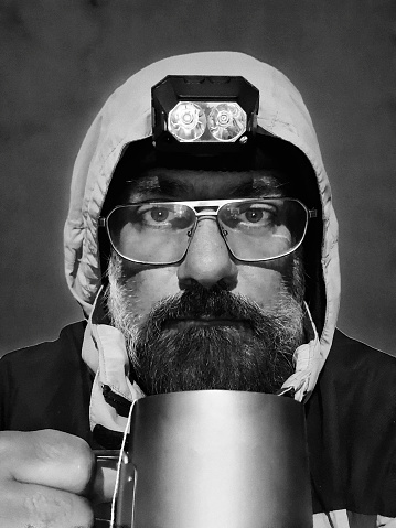 Portrait of a middle aged man with a tired expression holding a metal mug and wearing an explorer coat with a headlamp