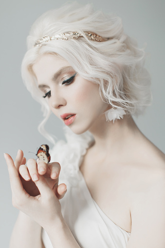 Young blonde model in studio. Woman with beautiful luxurious greek hairstyle in white dress. Girl with butterfly on hand. Golden wreath. Albino. Beauty portrait. Greek goddess. Purity and innocence