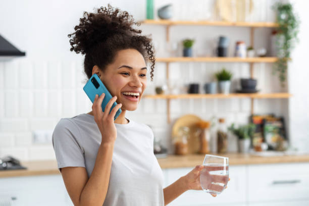 Smiling African American woman holding glass of clean water, talking on mobile phone while standing in kitchen at home, healthy lifestyle concept stock photo