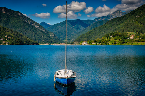 Sailing boat with white sail on Lake Garda, Italy.  Green water and trees in the foreground, Monte Baldo in the background