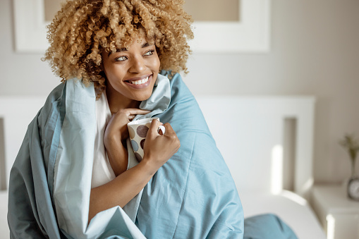 Pretty Woman Snuggled Under Duvet With Cup of Coffee or Tea in Bed at Home Bedroom. Morning, Leisure and People Concept