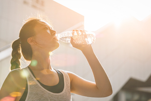 Young Asian woman wearing a white vest in the sunlight outdoors, drinking water to rest after exercising.