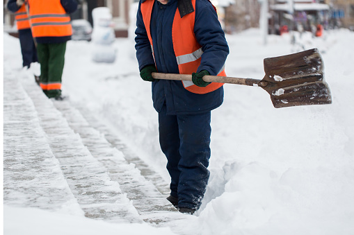 Employees of municipal services in a special form are clearing snow from the sidewalk with a shovel.