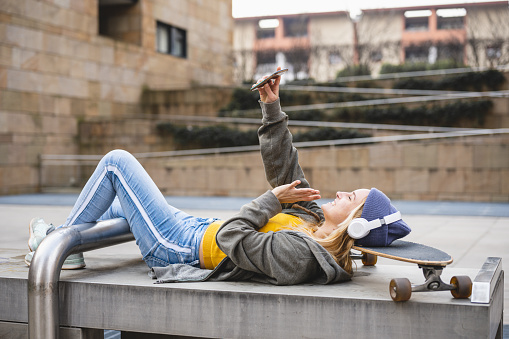 Young woman lying down with head on skate, holding smart phone to have a call, listening music and enjoying contents on line