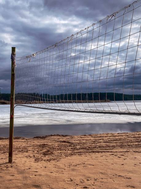 Deserted beach with volleyball net, lake covered with ice, cloudy moody sky over water, first spring days on the beach, sports