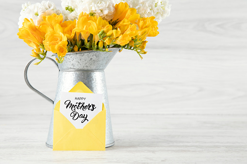 Happy Mother's Day with white and yellow flowers in a vase with greeting card in yellow envelope on rustic wooden background.