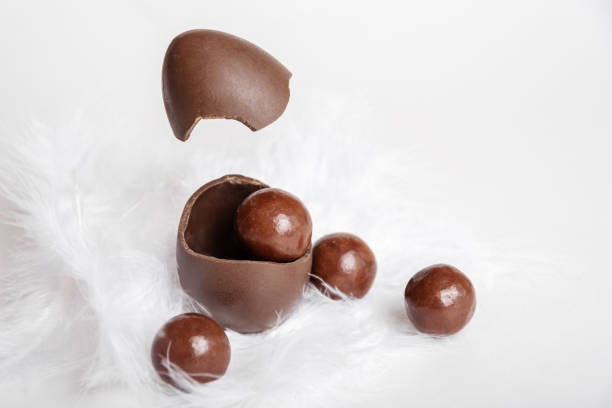 Cracked chocolate easter egg with chocolate small round candies and flying eggshell in white feather nest, copy space, levitation. Chocolate treat for kids. Easter concept.