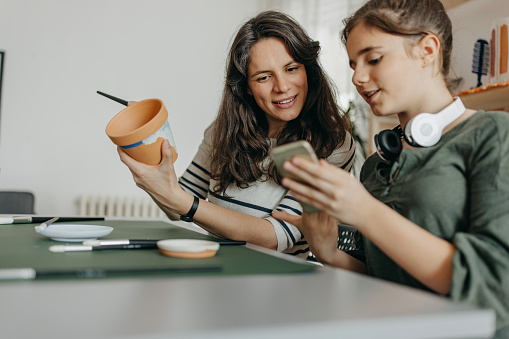 Creative mother and teenage daughter painting  ceramic pottery at home and using phone