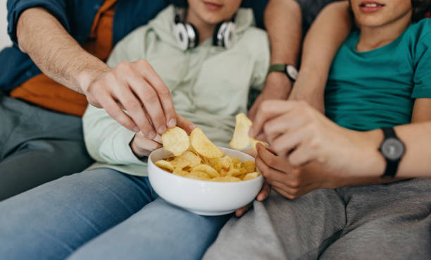 Taking a snacks Happy and smiling family sitting together on sofa at home, eating potato chips and watching tv family eating potato chips stock pictures, royalty-free photos & images