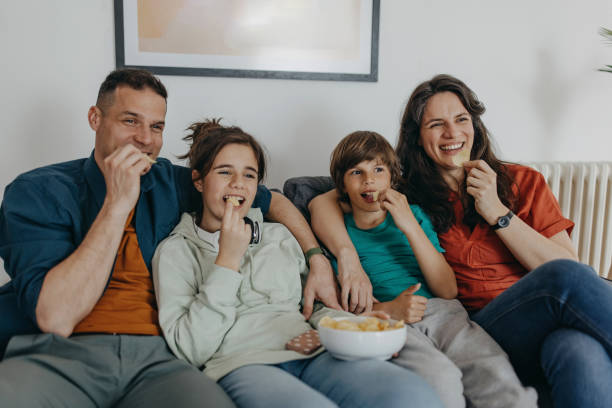 We love that movie Smiling family sitting together on sofa at home and watching funny movie on tv family eating potato chips stock pictures, royalty-free photos & images